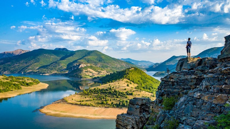 A solitary hiker stands on a rocky cliff overlooking a peaceful mountain lake under a blue sky., tags: und natürliche schönheit - pexel